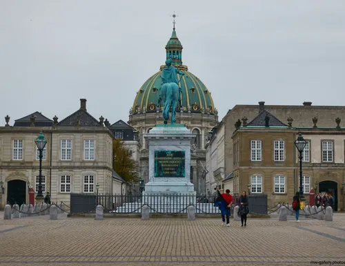 This urban architecture photograph, taken in the afternoon during the autumn season, shows Amalienborg Palace Square in the city of Copenhagen. The main subject is the equestrian statue of Frederick V, prominently positioned at the center of the square. Behind the statue, the dome of the Marble Church (Frederik's Church) can be seen, serving as a well-known landmark in Copenhagen. The image also features classic city architecture with the surrounding buildings of Amalienborg, which are notable for their historical significance in Denmark's capital. Several people are visible walking across the square, contributing to the active urban atmosphere of this area in Copenhagen.